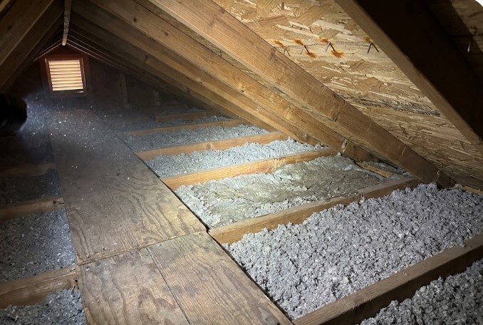 Attic with gray blown-in insulation between the joists, a plank walkway, and rust-stained roofing nail tips poking through the underside of the roof decking.