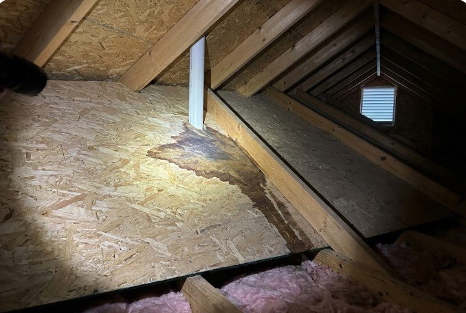 Flashlight beam on a dark water stain spreading across OSB boards around a white PVC plumbing vent pipe in an attic, with pink fiberglass insulation between the joists and a gable vent at the far end.